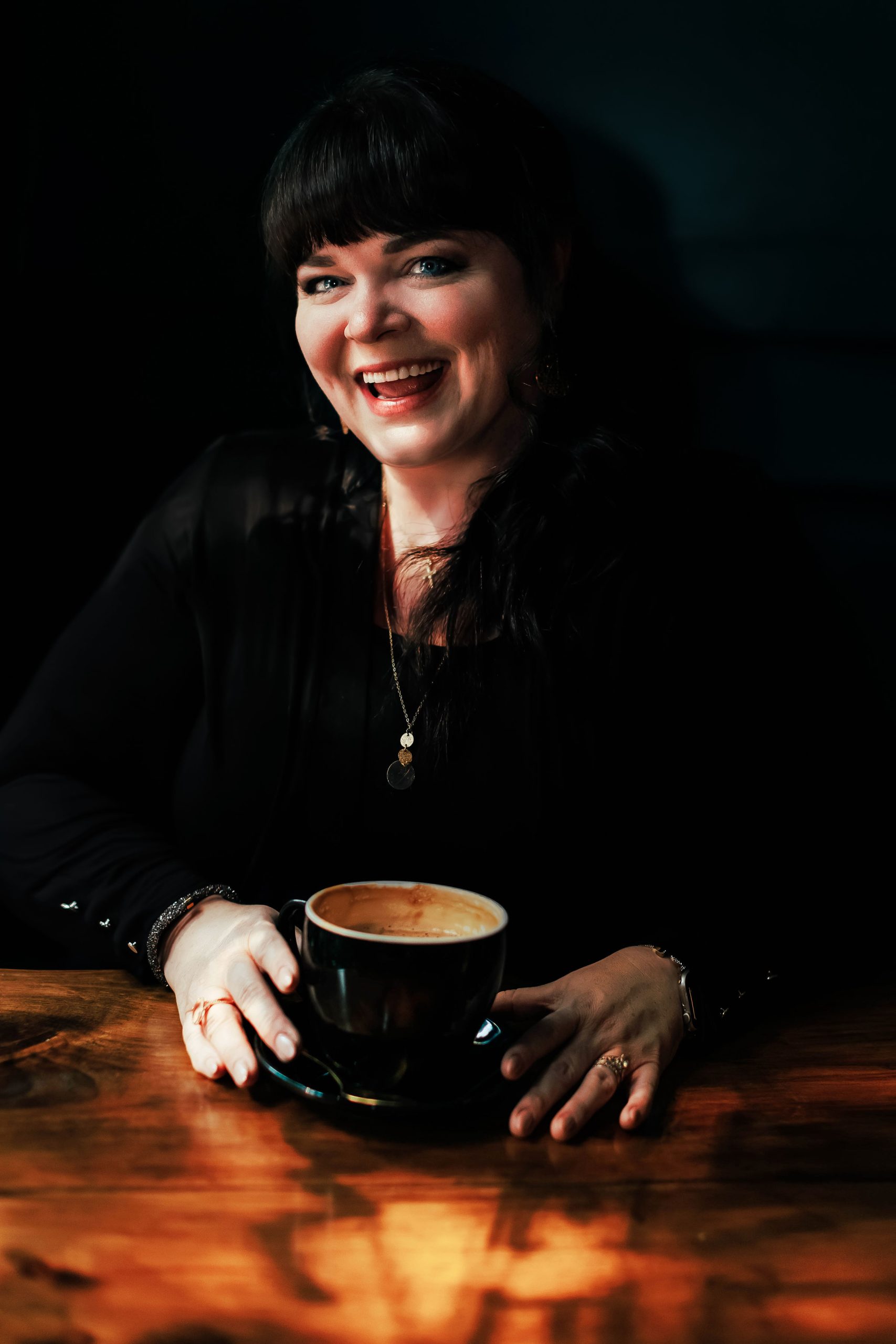 Jenn Henry smiling brightly with a coffee in hand, seated at a warmly lit table—reflecting approachability, creativity, and confidence.