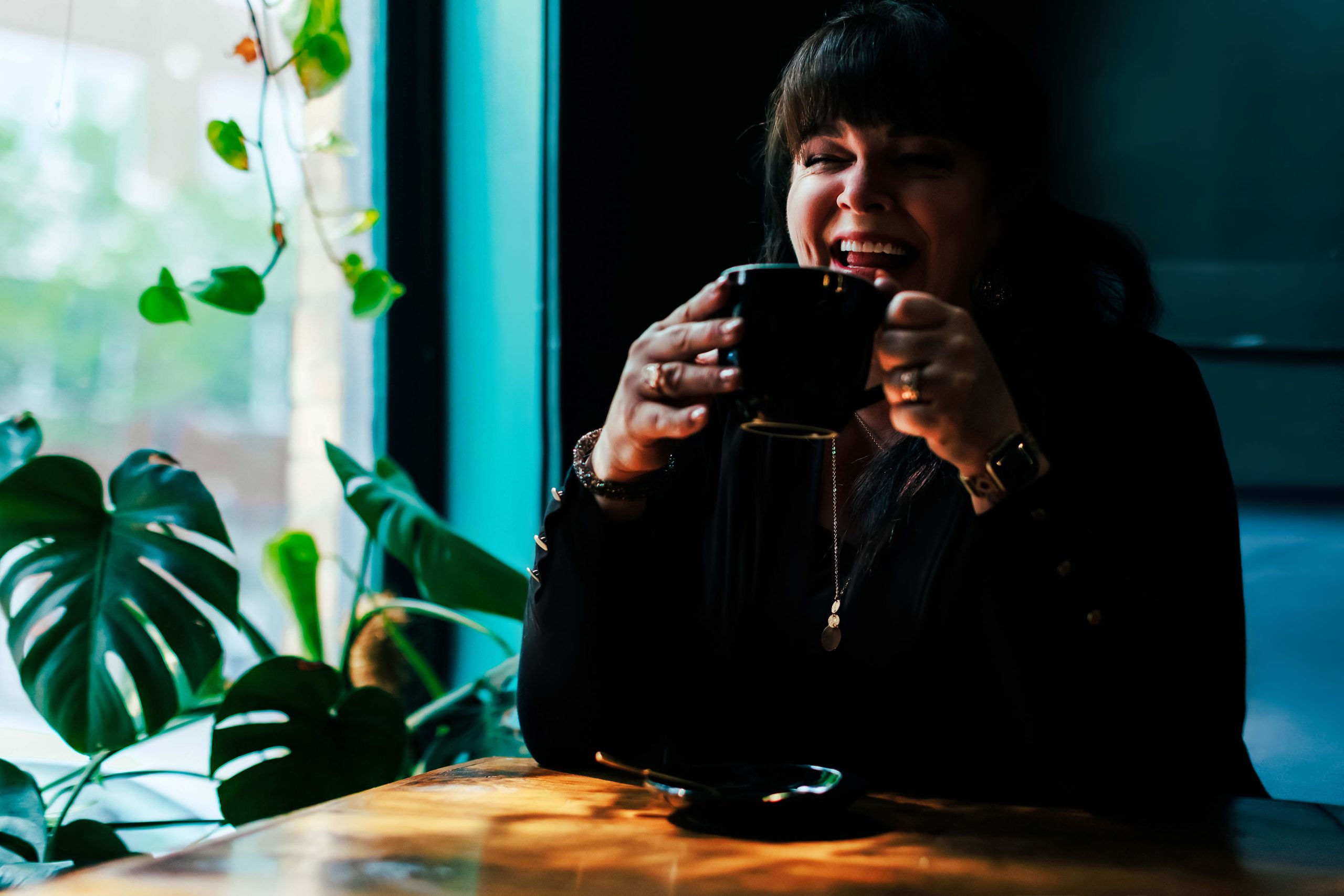 Jenn Henry smiling and holding a coffee mug while seated at a sunlit café table, with vibrant green plants and natural light streaming through a nearby window.