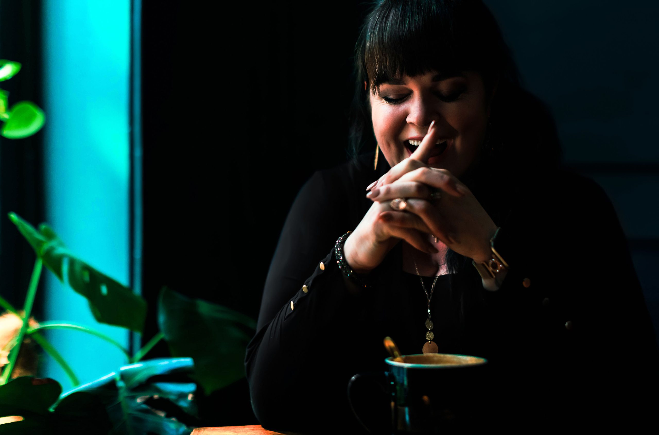 Jennifer Henry “Jenn Henry smiling with hands clasped in a quiet, joyful moment at a café table—surrounded by warm coffee and lush green plants.